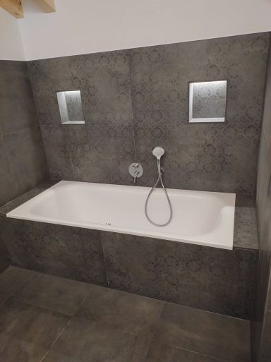 Badewannenbereich mit dunklen Fliesen und zwei beleuchteten Wandnischen
